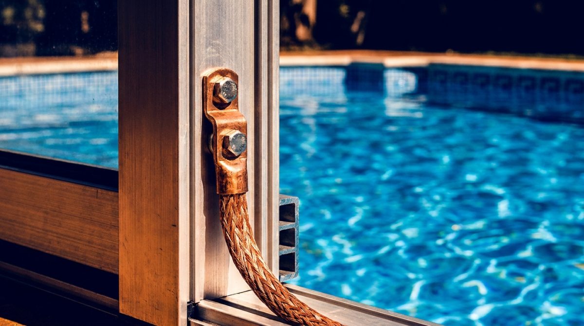 Close-up of an aluminium pool enclosure frame profile with a visible bonding lug and copper earthing conductor attached, pool water visible in the background