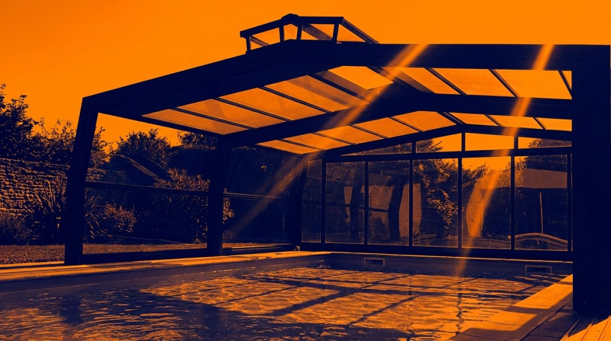 An aluminium pool enclosure on a sunny UK garden day with sunlight streaming through the glazed roof panels onto a pool below, ridge vent visible at the apex