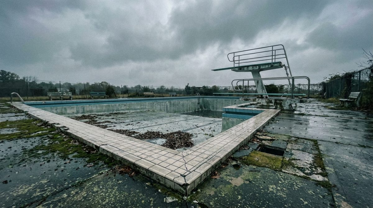 An ageing outdoor public swimming pool with cracked coping tiles and weathered concrete surrounds, viewed from the end of the pool on a grey overcast day